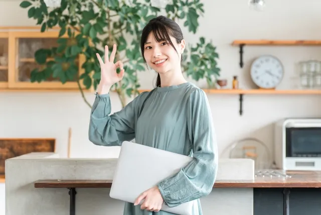 白いノートパソコンを左の小脇に持ち笑顔の女性が右手でOKサインをしている