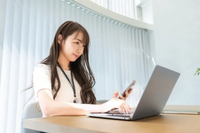 若い女性がテーブルでスマホを見ながらノートパソコンを操作している様子