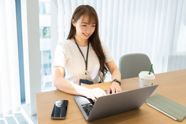 テーブルで白い服を着た女性がノートパソコンを操作していて、その近くにはスマホとタブレットとストローのついた飲み物が置かれている