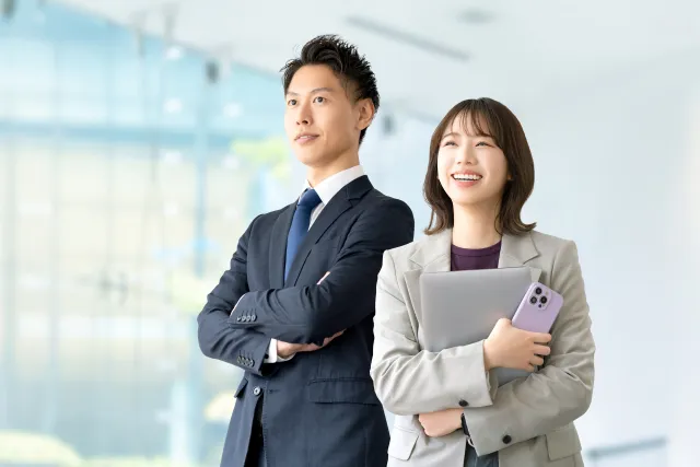 スーツをきた若い男性とジャケットを着たノートパソコンとスマホを持っている若い女性が笑顔で立っている