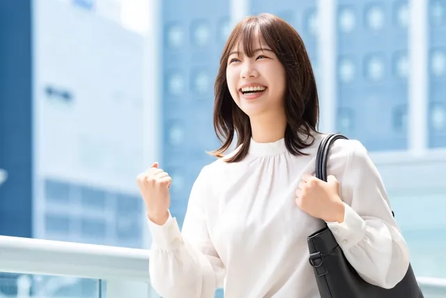 ビルをバックにして左肩にバッグを持ち右手でガッツポーズしている笑顔の女性