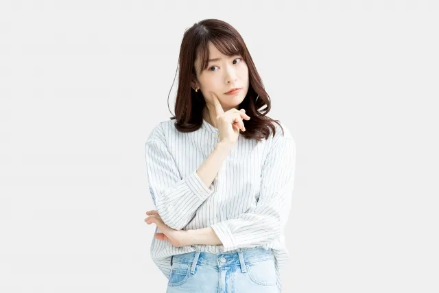 右手の人差し指を頬に当てて考えているストライプのシャツにジーンズ姿の女性
