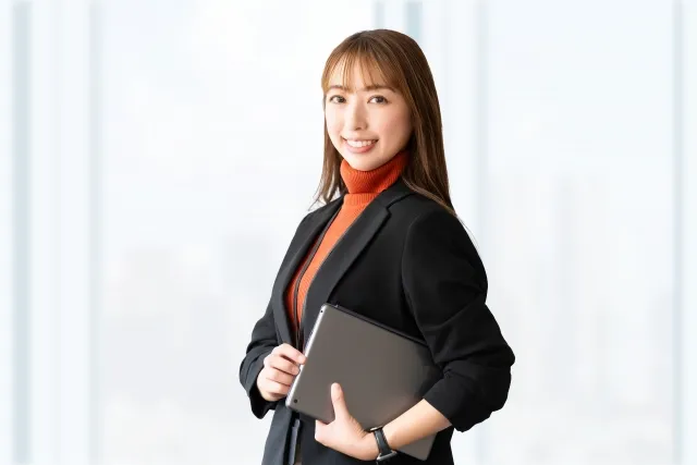 ノートパソコンを左の小脇に抱えて笑顔の黒いジャケットを着た女性