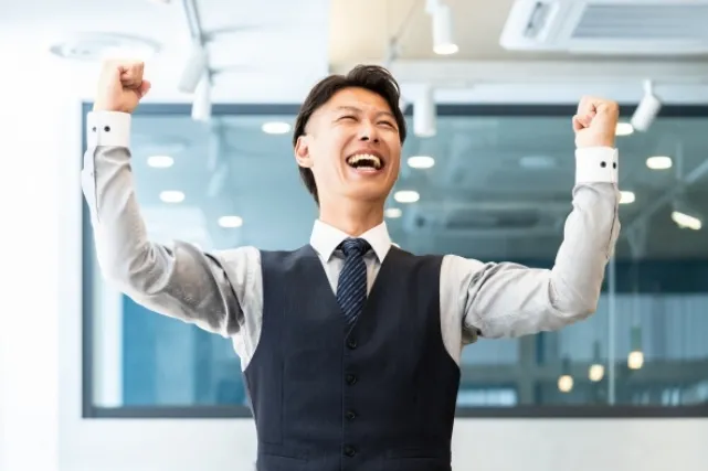 オフィスで立ち上がって両手でガッツポーズをする笑顔のスーツを着た男性
