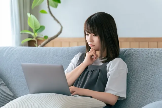 ソファに座って膝に乗せたクッションの上のパソコンを見ながら考えている女性