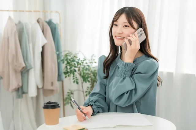 部屋のテーブルでスマホで会話しながら右手でメモをしている笑顔の女性