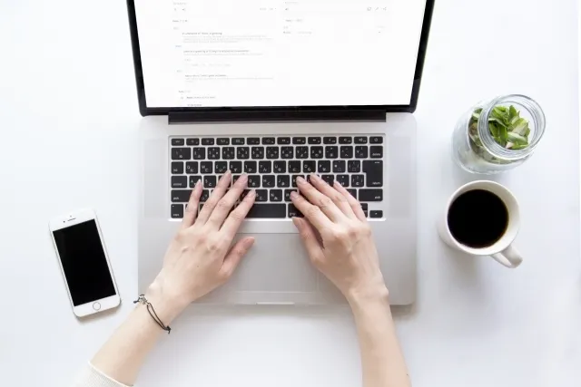 ノートパソコンを操作している両手と傍らにおいてあるスマホとコーヒーと置物の植物