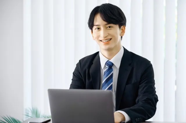 テーブルに置いたノートパソコンをいじりながらこちらを向いて笑顔のスーツを着た男性