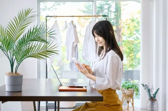 自宅のテーブルにノートパソコンと手帳を置いてスマホを見ている髪の長い女性