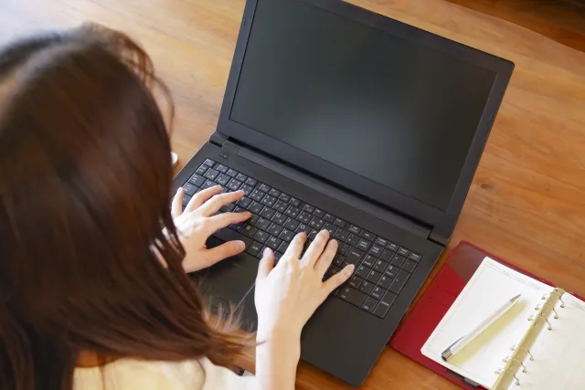 テーブルで黒いノートパソコンを両手で操作している髪の長い女性を斜め上の後ろから見ている様子