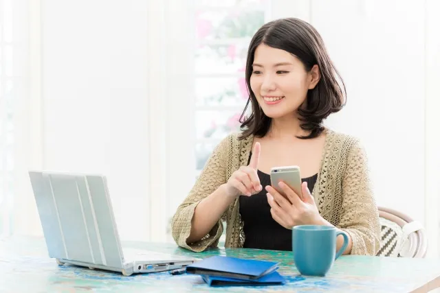 テーブルでノートパソコンを見ながらスマホを操作しているカーディガンを着た女性