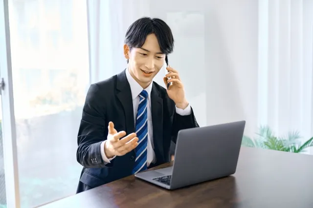 スーツ姿の男性がノートパソコンを見ながらスマホで電話している