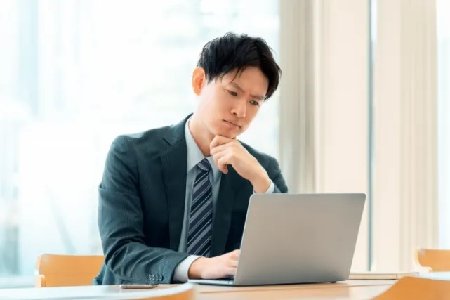 デスクでノートパソコンを見ながら左手で頬杖をして考えているスーツを着た男性