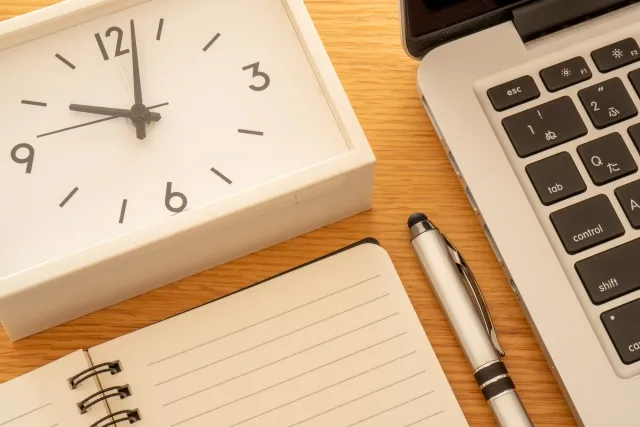 ノートパソコンの横に置かれた白色の四角い置時計とノートとボールペン